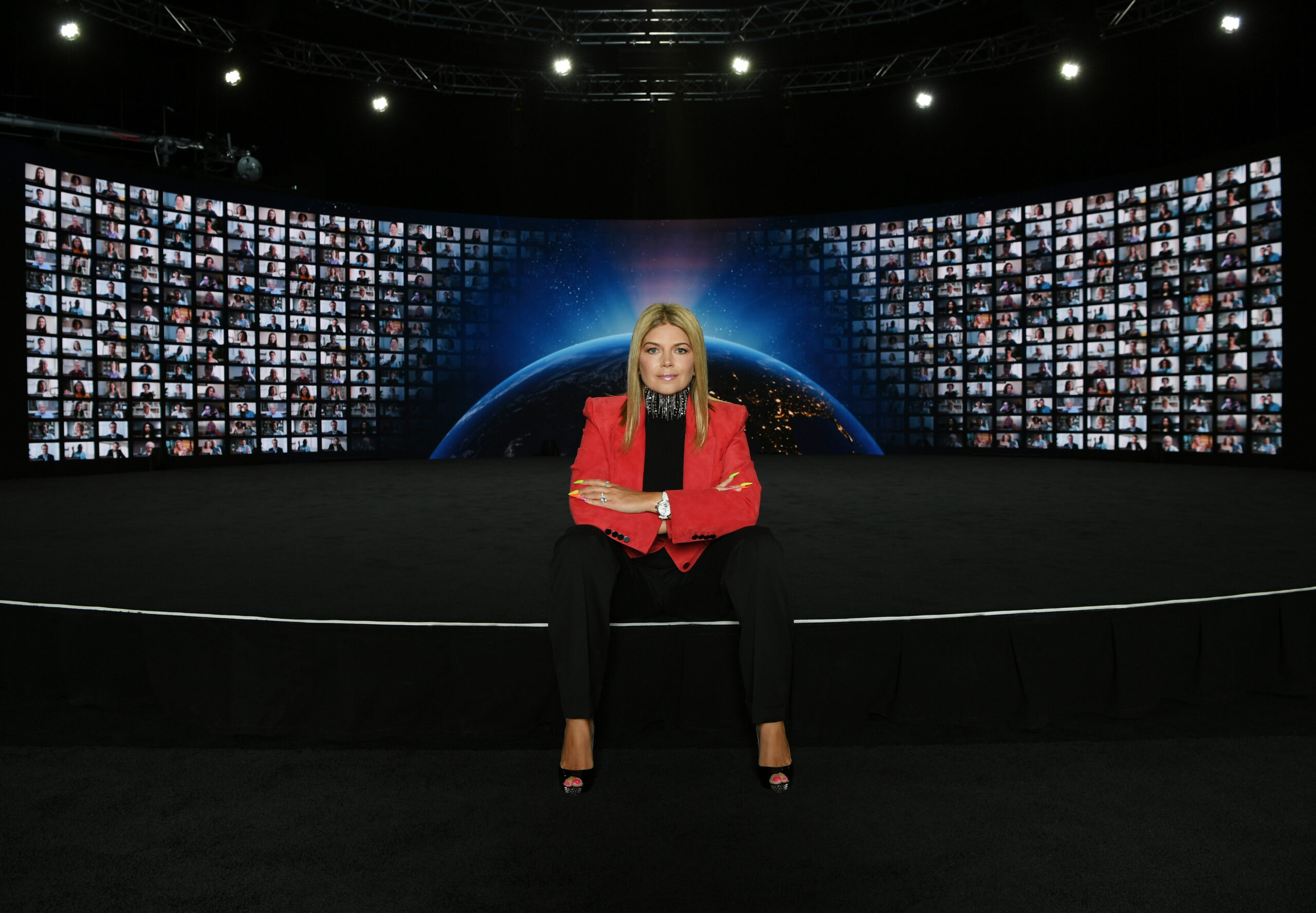 Marina Worre sitting in front of a stage with red blazer behind her a video wall with participants to her event.